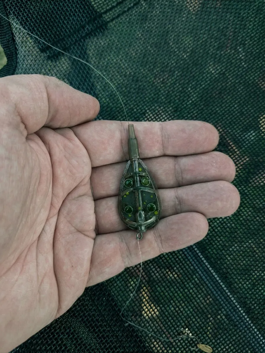 Een method feeder van 20 gram met een onderlijn in de hand, boven een groene mat