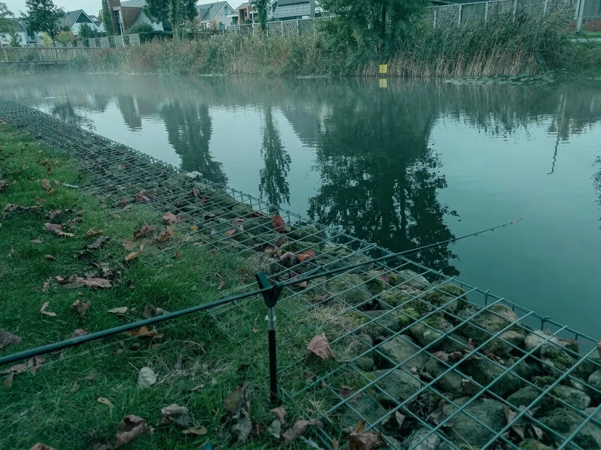 De Stakenbeek in Oldenzaal met een feederhengel op de steun, een beschoeiing van stenen in gaas en een stille waterspiegel met weerspiegelde bomen